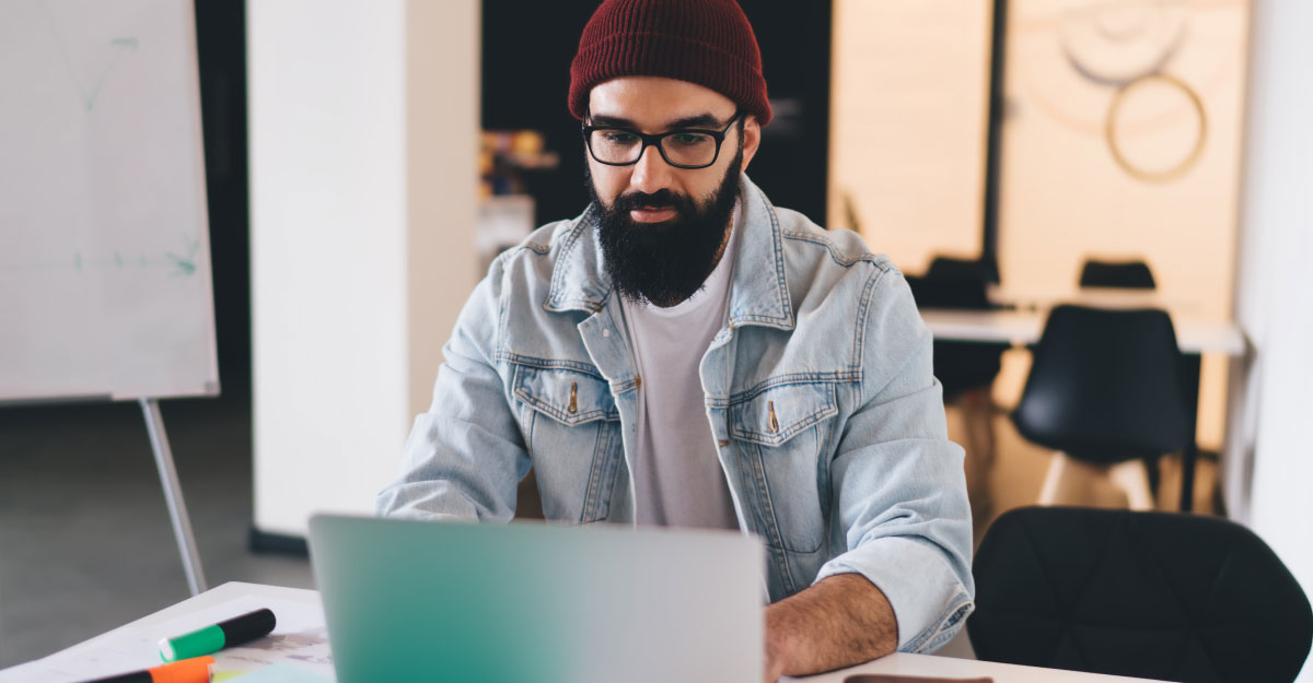 A man with a dark beard, glasses, and knit beanie works on a laptop in a modern office.