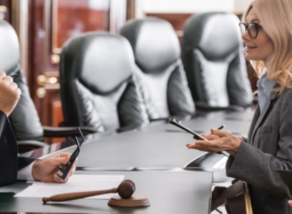 A female lawyer with blonde hair speaks with a gray-haired male judge in a legal court. A gavel sits on the table between them.