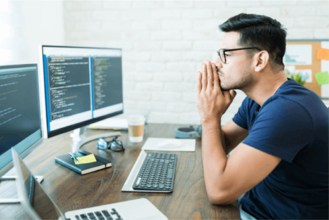 A man is intensely focused on his work at a computer desk. He is sitting in front of two large monitors displaying code. He is wearing glasses and has his hands clasped together in front of his face, appearing deep in thought. The desk is organized with a notebook, a keyboard, and a cup of coffee. In the background, there is a bulletin board with sticky notes and a few plants.