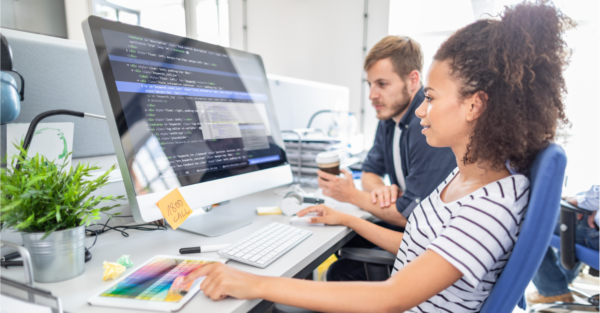 A young woman with curly hair writes lines of code on a desktop computer, while a young man looks on beside her.