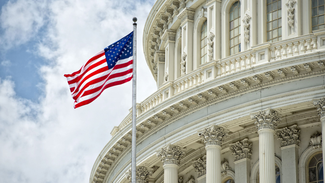 The image shows the American flag waving in front of the U.S. Capitol building with ornate columns and arched windows, set against a partly cloudy sky.